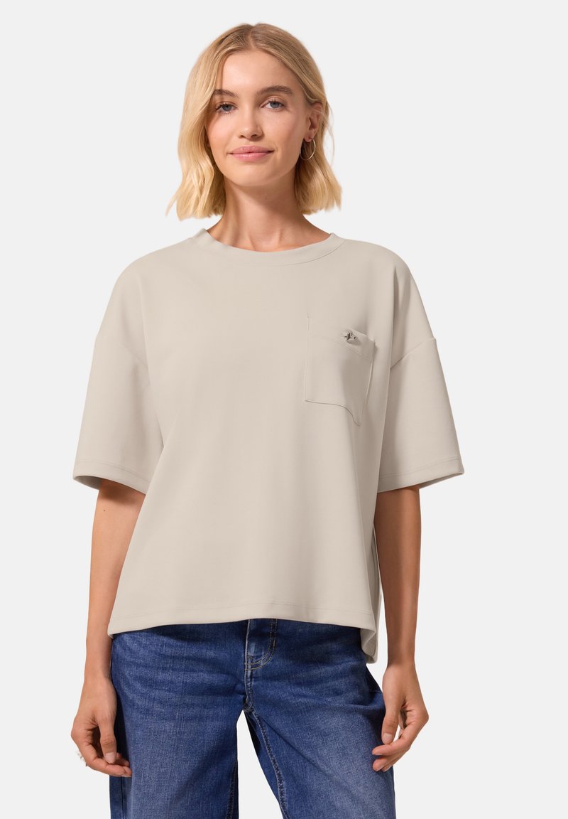 Femme blonde portant une chemise beige oversize à manches courtes avec une poche poitrine et un jean bleu, debout devant un fond uni.