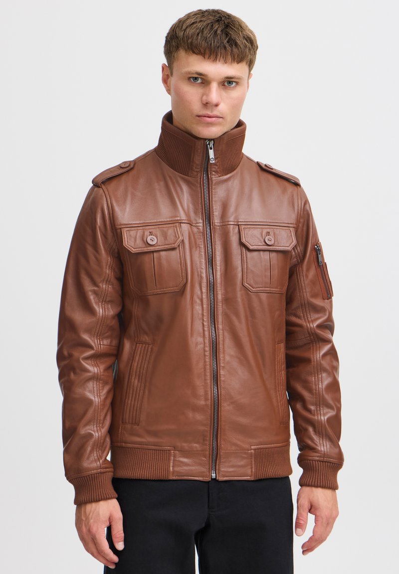 Veste en cuir marron avec col montant, deux poches poitrine et poignets côtelés. Dotée d'une fermeture éclair à l'avant et d'épaulettes. Texture lisse.