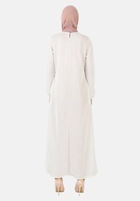 Lang, hvid kjole med lange ærmer og en nøglehulldetalje i ryggen, parret med en lys pink hijab og nude stilethæle. Blødt, flydende stof.