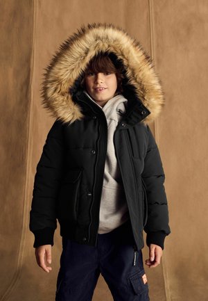 Schwarze Pufferjacke mit einer mit Fell gefütterten Kapuze, ausgestattet mit einem Reißverschluss und mehreren Taschen. Getragen über einem grauen Hoodie und navyblauen Cargo-Hosen.