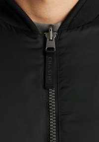 Close-up van een zwarte jasrits die gedeeltelijk is dichtgeritst over een grijs shirt, waarbij details van de kraag en de ritstrekkertje zichtbaar zijn.