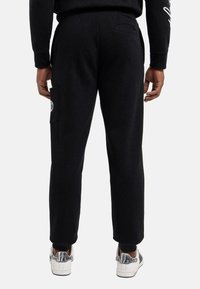 Svarte joggebukser med oppbrettede ankler, har et sidelommer med logo-lapp, og en glatt bomullstekstur. Brukt med hvite sneakers.