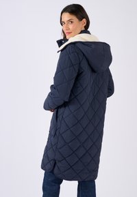 Gequilted marineblauw jack met een faux fur gevoerde capuchon, met een middellange ontwerp en zijzakken. Gestructureerd oppervlak met diamanten patronen.