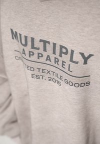 Grå sweatshirt gjord av mjukt tyg; har djärv svart text "MULTIPLY APPAREL" och "CRAFTED TEXTILE GOODS EST. 2015" i grafisk design.