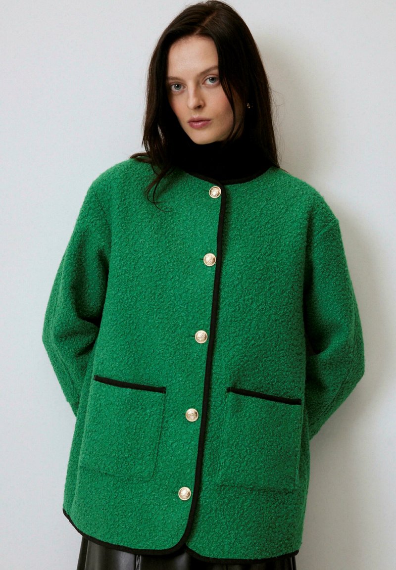 Jachetă de fleece verde cu detalii negre, având un decolteu rotund, buzunare mari în față și butoane aurii proeminente. Material moale, texturat.