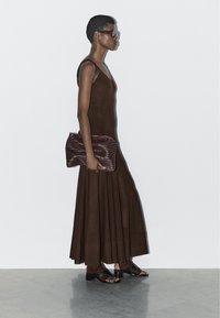 Brązowa sukienka bez rękawów z plisami, teksturowana ciemnoczerwona kopertówka oraz czarne sandały na obcasie typu chunky. Modelka pozuje na jednolitym tle.
