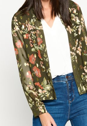 Kvinde iført en olivengrøn gennemsigtig blomstret bluse med gyldne accenter, hvid v-hals top og blå jeans med høj talje.