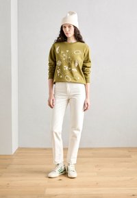Sweatshirt vert olive avec broderie florale blanche, associé à un jean blanc droit et des baskets crème avec des rayures vertes. Bonnet en tricot blanc.