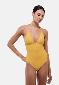 Maillot de bain jaune à col halter en tissu texturé, avec des bords festonnés au niveau du décolleté et une silhouette ajustée qui met en valeur la forme du corps.