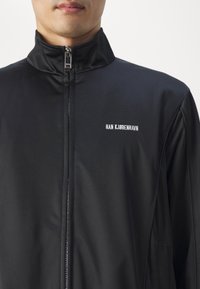 Black zip-up jacket made from smooth fabric. Features a high collar and logo "HAN KJØBENHAVN" in white on the upper left.