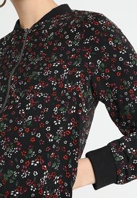 Veste noire à manches longues avec imprimé floral petit en rouge, violet, blanc et vert, fermeture éclair à l'avant, col et poignets côtelés.