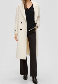 Dubbelzijdige beige jas van textuurstof, met zwarte knopen, twee voorkezen, gelaagd over een zwarte top en zwarte broek.