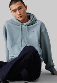 Felpa azzurra chiara con cappuccio e cordino, morbida al tatto, piccolo logo ricamato sul petto, abbinata a jeans scuri.