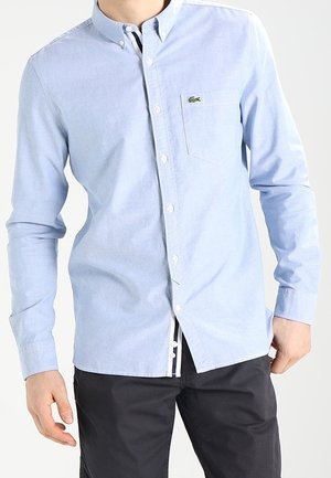 Uomo che indossa una camicia azzurra chiara a maniche lunghe con taschino sul petto e pantaloni scuri, in piedi contro uno sfondo semplice.