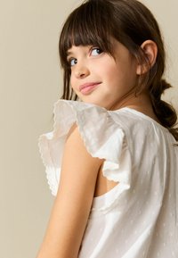 Balts kokvilnas tops ar apaļu kakla izgriezumu un volānu piedurknēm ar smalkām viļņotām malām un teksturētu punktu rakstu, redzams no sāniem.