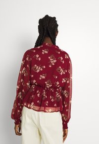 Kvinna med flätat hår som bär en röd blommig långärmad blus och vita byxor, sedd bakifrån mot en vit bakgrund.