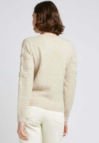 Pull en maille beige avec des accents floraux texturés sur les manches, un col rond, et des poignets et un ourlet côtelés. Associé à un pantalon de couleur claire.