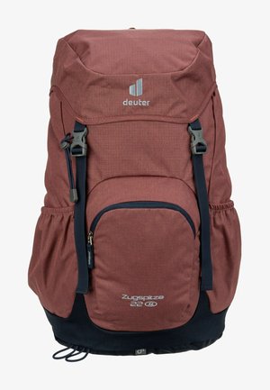Mochila de tela roja con solapa superior, bolsillo frontal con cremallera, bolsillos de malla en los laterales, correas ajustables y panel inferior azul oscuro. Presenta logo.