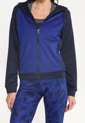 Woman wearing a blue and black zip-up hoodie over a gray top and patterned blue leggings standing against a plain background.