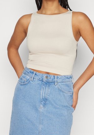 Kobieta w beżowym, bezrękawnikowym krótkim topie i jasnoniebieskiej dżinsowej spódnicy z wysokim stanem, z rękami swobodnie opuszczonymi wzdłuż ciała.