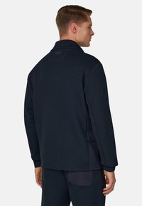 Giacca pullover blu navy scuro, realizzata in tessuto morbido, con colletto alto, tasche laterali e vestibilità comoda. Dettaglio testurizzato sulla spalla visibile.