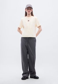 Εκρού κοντομάνικη μπλούζα με μικρό λογότυπο, συνδυασμένη με γκρι παντελόνι με ρίγες και φαρδιά πόδια και μαύρα παπούτσια, συνοδευόμενη από γκρι καπέλο.