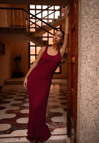 Geribbelde, getailleerde bordeauxrode jurk met een ronde halslijn en een geschulpte zoom. Op de achtergrond is een trap te zien en een betegelde vloer met cirkelvormige patronen.