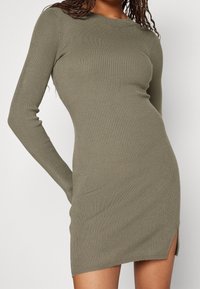 Olivgrön ribbad stickad klänning med långa ärmar, rundad halsringning och sidoklyfta för lätt rörlighet. Mjuk textur och figursydd silhuett.