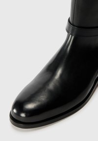 Botină de piele neagră până la gleznă cu vârf ascuțit, textură netedă, cusături minime și talpă rotunjită cu o strălucire subtile.