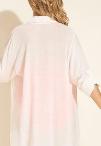 Blondyn ubrany w luźną, długą białą koszulę z podwiniętymi rękawami, widziany z tyłu na tle jednolitego, jasnego tła.