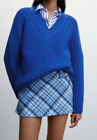 Femme portant un gros pull en maille bleu sur une chemise rayée bleu et blanc, assorti d'une mini-jupe à carreaux bleus.
