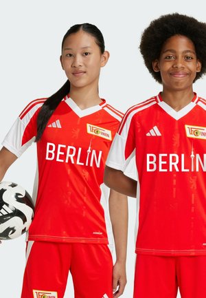 Magliette di calcio rosse e bianche con "BERLIN" stampato in caratteri grandi. Presentano il marchio Adidas e un design stilizzato. Giocatori che tengono in mano un pallone da calcio.