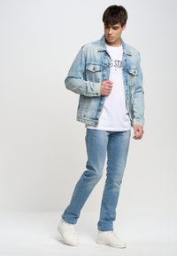 Chaqueta de denim azul claro con dos bolsillos en el pecho, llevada sobre una camiseta gráfica blanca, combinada con jeans azul claro y zapatillas blancas.