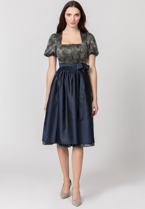 Frau trägt ein knielanges Kleid mit Puffärmeln und Blumenmuster, kombiniert mit einer marineblauen Schürze, die in der Taille mit einer Schleife gebunden ist.