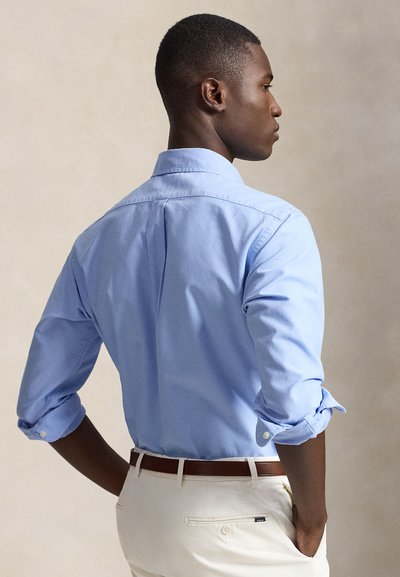 Homme portant une chemise bleu clair à manches longues avec les manches retroussées, pantalon beige et ceinture marron, debout les mains dans les poches.