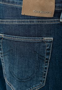 Poche arrière d'un jean en denim bleu avec une pièce en cuir portant l'étiquette « True Religion » et des détails de couture orange visibles.