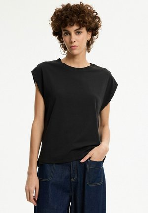 Mujer con cabello rizado que lleva pendientes de aro, camiseta negra sin mangas y vaqueros azul oscuro, con una mano en el bolsillo, de pie frente a un fondo blanco.
