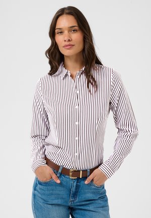 Femme aux longs cheveux foncés portant une chemise blanche à fines rayures verticales foncées et un jean bleu avec une ceinture marron, les mains dans les poches.