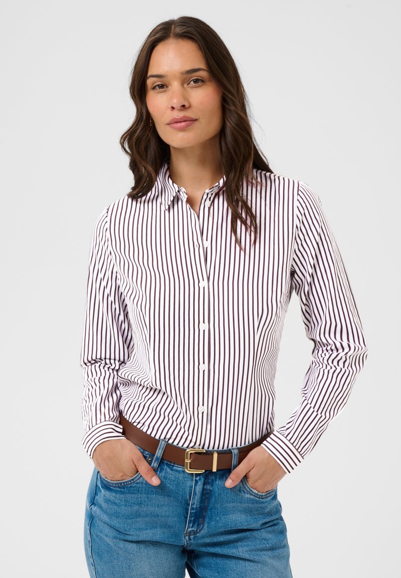 Femme aux longs cheveux foncés portant une chemise blanche à fines rayures verticales foncées et un jean bleu avec une ceinture marron, les mains dans les poches.