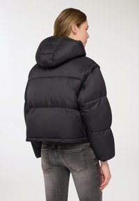 Giacca imbottita cropped nera con cappuccio, caratterizzata da un design trapuntato e una texture liscia. Abbinata a jeans grigi, che mettono in risalto una silhouette aderente.