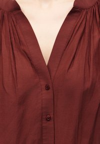 Κοντινό πλάνο ενός μπλουζιού με λαιμόκοψη σε σχήμα V σε σκουριά απόχρωση, με κουμπιά μπροστά και λεπτομέρεια με ραμμένο ύφασμα στους ώμους.