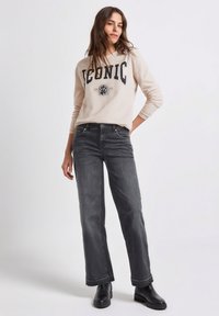 Felpa beige chiaro con la scritta "ICONIC", abbinata a jeans a gamba larga grigio scuro e stivaletti neri; vestibilità rilassata e tessuto morbido.