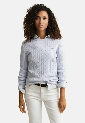 Kobieta o kręconych włosach, ubrana w jasnoniebieski sweter z grubym warkoczowym splotem na białej koszuli z kołnierzykiem oraz białe spodnie z czarnym paskiem, stojąca nieruchomo.