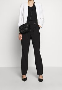 Femme portant un blazer blanc, un haut noir, un pantalon noir taille haute, des talons pointus noirs et un sac banane matelassé noir.