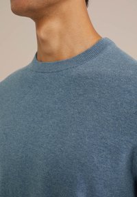 Blå stickad tröja med rund halsringning, med en mjuk textur och subtila färgvariationer. Närbild på tyget.