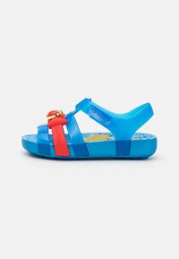 Blå gjennomsiktig plast sandal med en rød stropp og dekorativt hardware. Teksturert blå såle med et fargerikt mønstrete fotbed.