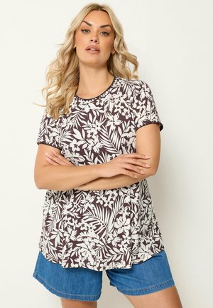 Blond kvinde med bølget hår iført en brun og hvid blomsterkortærmet top og blå denimshorts, arme krydset.