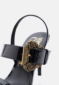 Μαύρο δερμάτινο σανδάλι με ψηλό τακούνι και χρυσό λουλούδι αγκράφα με χαραγμένη την επιγραφή "Versace Jeans Couture" στο λουράκι του αστραγάλου.