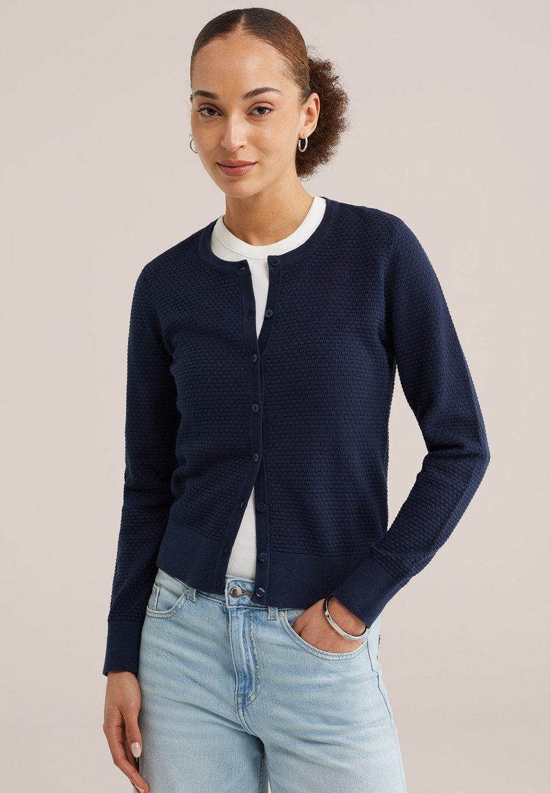 Femeie purtând un cardigan albastru închis texturat, cu nasturi, peste un top alb, blugi deschiși la culoare, cu mâna dreaptă în buzunar, pe un fundal bej.