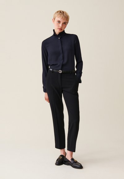 Blusa navy con collo a volant e maniche lunghe; abbinata a pantaloni neri su misura. Il modello indossa mocassini neri con dettagli dorati.